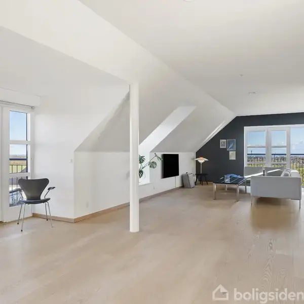 Lyst stueområde med grå sofaer og stol omkring sofabord. Store vinduer og en dør giver udsigt til balkon og natur. En lampe og grøn plante supplerer rummet.