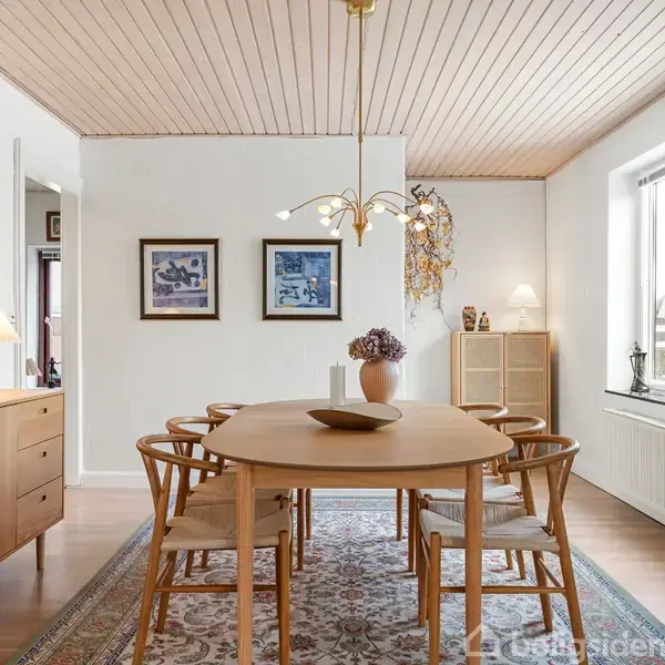 Ovalt træbord med otte stole står på orientalsk tæppe. Loftslampe hænger over bordet. Skænk med lampe til venstre, malerier på væggen, stort vindue til højre.