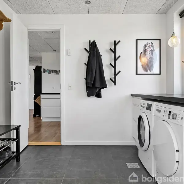 En moderne køkkenø med hvid front og sort bordplade. En sort emhætte hænger over grønne fliser. I baggrunden ses spisebordsstole på et trægulv, med billeder på væggen.