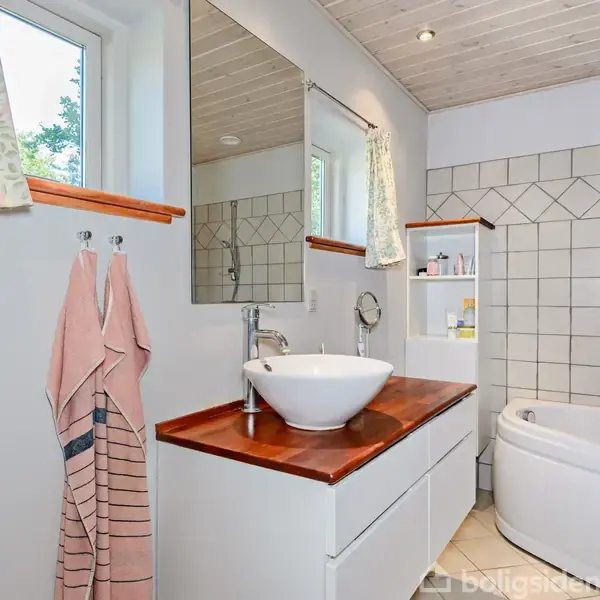 Hvid vask står på en træbordplade, omgivet af fliser og badekar i et lyst badeværelse med vindue og håndklæder på knager.