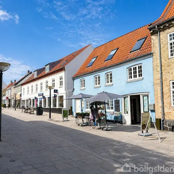En række farverige bygninger står langs en brolagt gågade. Folk sidder ved udendørs caféborde under parasoller, mens vejrudsigten er solrig og himlen er blå med få skyer.