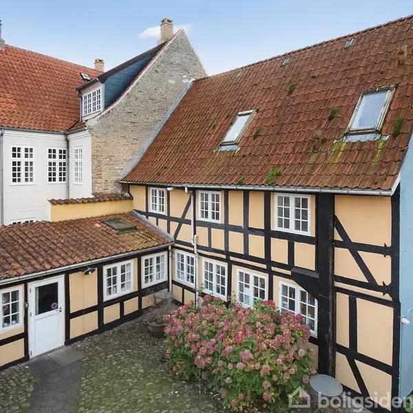 Et gammelt halv-timber hus står i en gårdhave, omgivet af blomster og brosten. Baggrunden viser nabohuse med røde tage og hvide vinduesrammer.