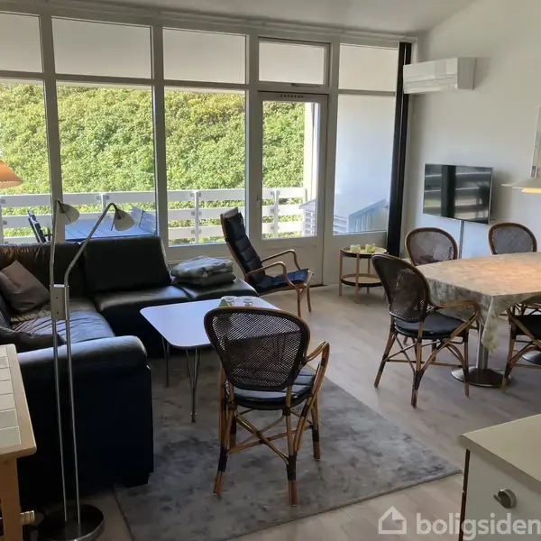 En stue med sort lædersofa og sofabord er udstyret med spisebord og stole i flet. Udsigt til terrasse og grønne træer udenfor vinduerne. En lampe hænger over bordet.
