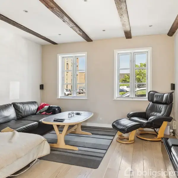 Sort lædersofa og lænestol står i en stue med to vinduer, hvide vægge, og trægulv. Et sofabord står foran sofaen, og et tv hænger på væggen.