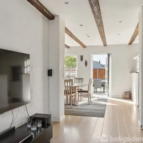 Fladskærms-TV hænger på en hvid væg i en stue, der leder til en spisestue med træbord og stole. Trægulve og synlige loftsbjælker skaber en hyggelig atmosfære.