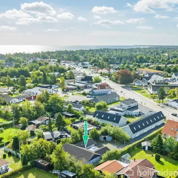 Hus indikeret med en grøn pil, beliggende i et grønt boligområde med spredte huse og veje. Baggrunden viser skov og kystlinje under en klar himmel med skyer.