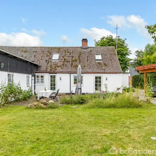 Et hvidt hus med skråt tag har en terrasse med havemøbler i en grøn have. En bil er parkeret under en carport til højre.