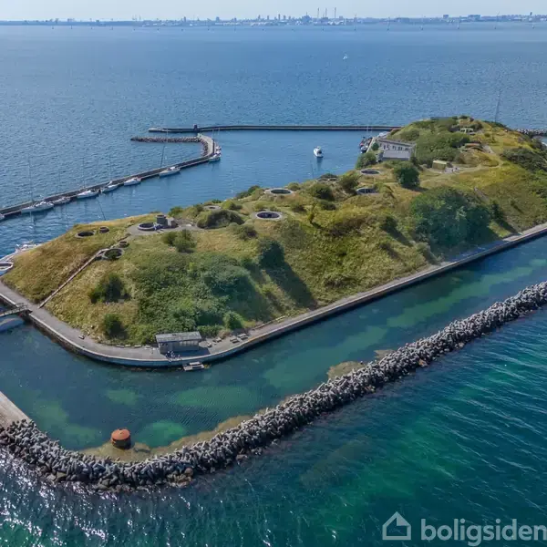 En lille, græsklædt ø omgivet af en stenmole ligger i havet. Øen har bygninger og stier, og byens skyline kan ses i horisonten.