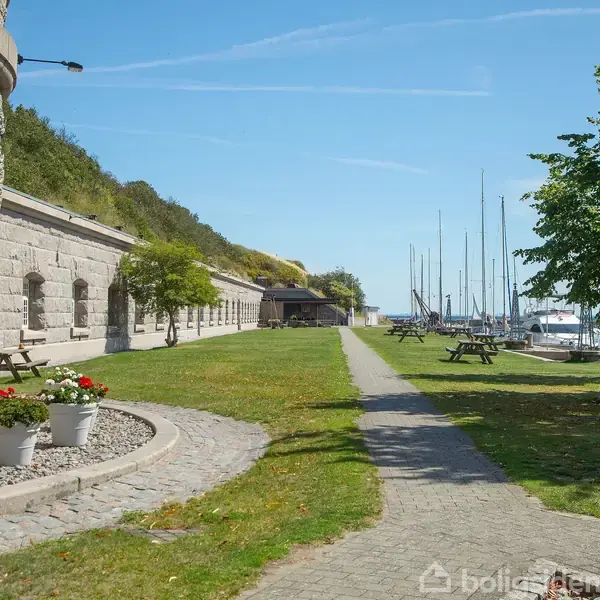 Stenbygning står langs en sti, der fører til en marina med sejlbåde. Græsplæne med picnicborde og træer skaber en afslappende atmosfære ved vandområdet.