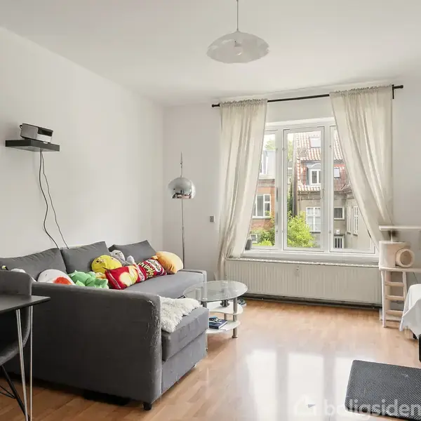 Stue med grå sofa fyldt med farverige puder, stående foran et stort vindue med hvide gardiner. En hængele lampe, spisebord og stol ses i rummet.
