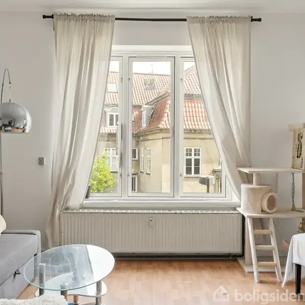 En stue med en grå sofa til venstre, dekoreret med farverige puder. Der står en rund glasbord foran den. En stor vindueskarm med gardiner trækker lyset ind. Ved siden af står et kattemøbel.
