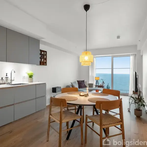 Et moderne køkken-alrum med spisebord og stole, er oplyst af en stor pendellampe. Et vindue viser havudsigt, mens en sofa og planter tilføjer hygge.