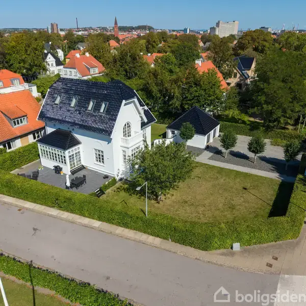 Hus med sort tag står i en villakvarter. Huset er omgivet af en hæk og har en terrasse med møbler. Omgivelserne har flere træer og andre huse.