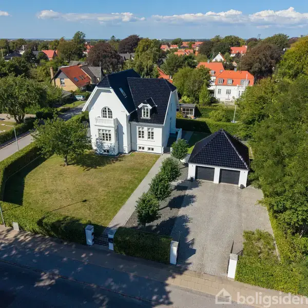 En hvid to-etagers villa med sort tag ligger på en velplejet græsplæne. Ved siden af er en garage med matchende tag. Omgivet af træer og buske i et boligområde.
