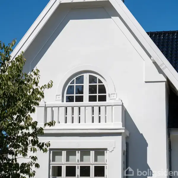 Et hvidt hus med en buet, vinduesdetalje over en lille balkon. En trægren med grønne blade ses i forgrunden.