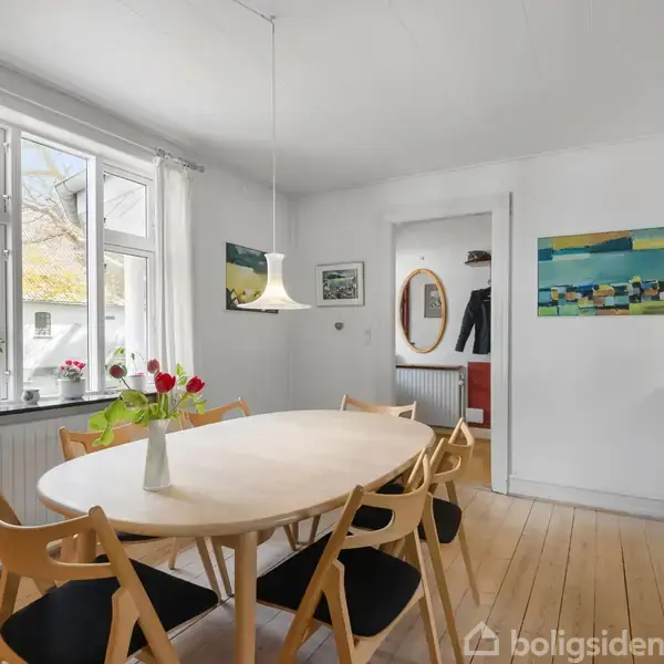 Et ovalt spisebord med seks stole står i et lyst rum. Væggene har kunstværker, og vinduet har hvide gardiner og planter på karmen. En lampe hænger over bordet.