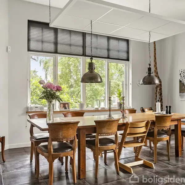 Spisebord med træstole omkring, dekoreret med en vase fyldt med blomster. To hængende lamper er over bordet. Et vindue med udsigt til grønne træer i baggrunden. Et ur hænger til venstre.