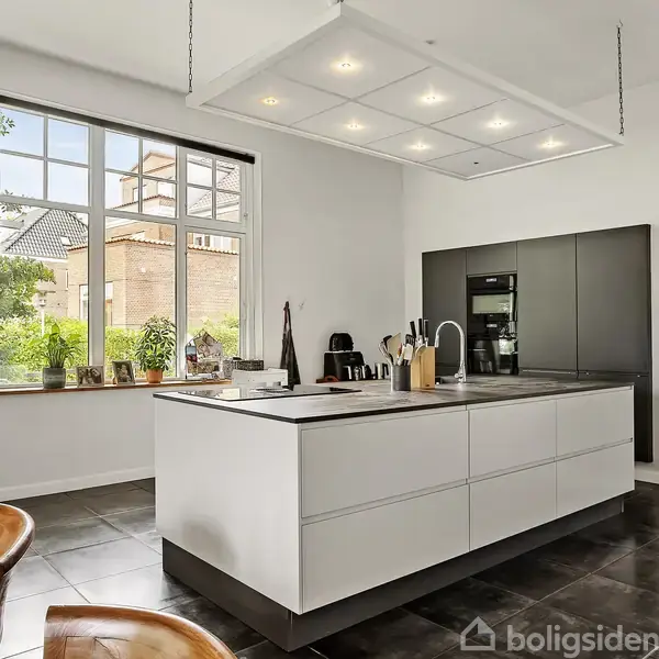 Køkkenø med hvide skuffer står i midten og er fyldt med køkkenredskaber. Store vinduer bagved lader naturligt lys strømme ind, og der er stilfulde træstole ved siden af.