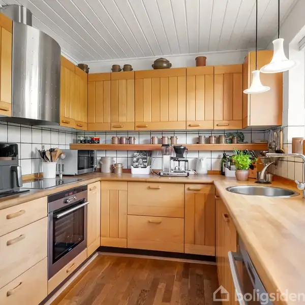 Et køkken med lyse træskabe, stålvask og keramisk komfur. Køkkenmaskiner er placeret på bordet, og planter står i vindueskarmen. Flisebelægning på væggen tilføjer dekorativt præg.