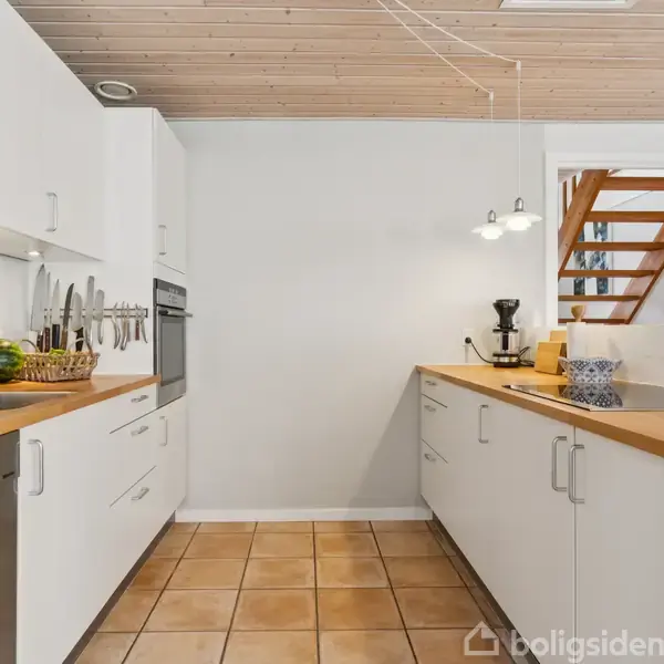 Et hvidt køkken med træbordplader indeholder en opvaskemaskine, vask, komfur og kaffemaskine. Køkkenet har flisegulv og træloft, med en trappe synlig i baggrunden.