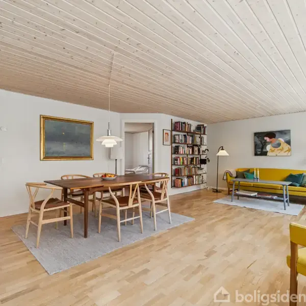 Spisebord med stole står på tæppe, omgivet af stue med sofa, bogreoler og malerier på væggen. Trægulv og træloft skaber en lys, rummelig atmosfære.