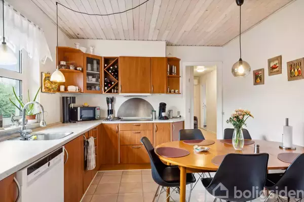 Et køkken med træskabe og en rund spisebord i forgrunden. Bordet har fire sorte stole og en vase med blomster. Lysekeramiske fliser og hvidt træloft udgør rummet.