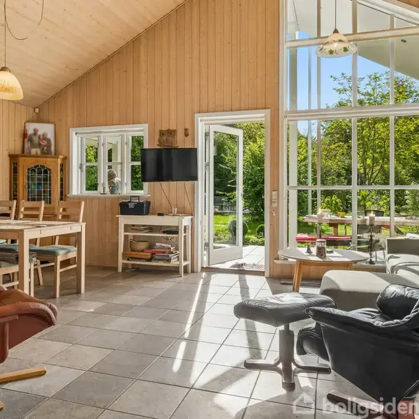 En hyggelig stue med lænestole og spisebord. Store vinduer og åbne døre giver udsigt til en grøn have. Lyse trægulve og vægge skaber en varm atmosfære.