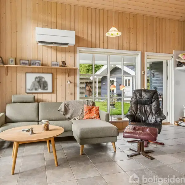 En stue med en grå sofa og lænestol står på flisegulv. Store vinduer slipper lys ind. Der er en brændeovn i hjørnet, et billede på væggen, og en dør fører ud til en have.