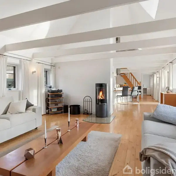 Sofaer står i en lys stue med trægulv og loftsbjælker. En moderne brændeovn er tændt, og spisebordet ses i baggrunden. Stuen har store vinduer med lyse gardiner.