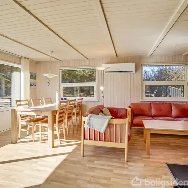 Stue med spisebord og seks stole, samt rød sofa og sofabord. Rummet har store vinduer med masser af naturligt lys. Lys træbeklædning og moderne belysning skaber en hyggelig atmosfære.