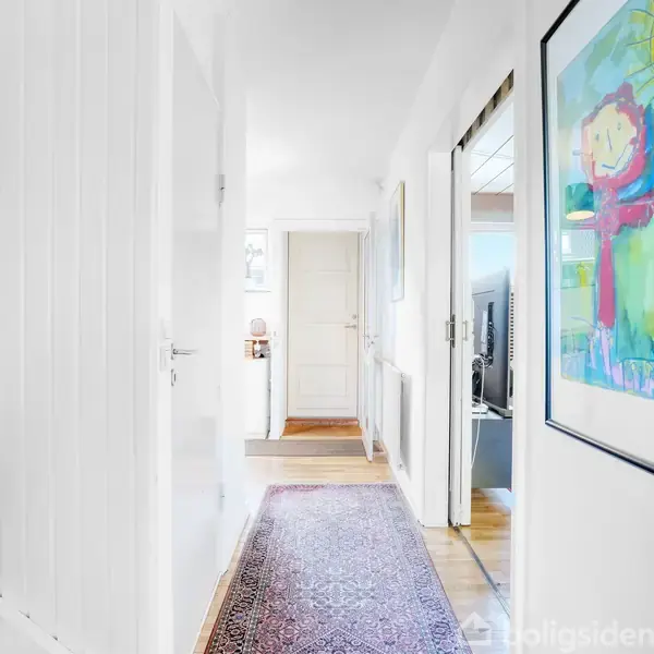 En smal gang med trætrappe til venstre og farverigt kunstværk på væggen. Et tæppe løber langs gulvet mod en dør for enden af gangen.