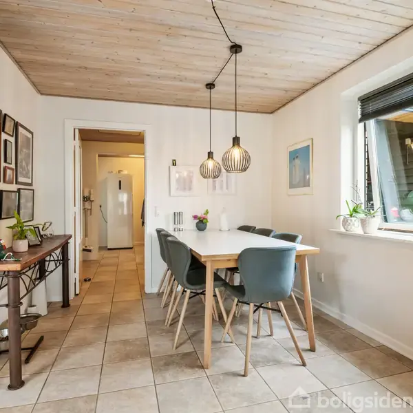 Et spisebord med otte stole står i et lyst rum. Over bordet hænger tre lamper. Et vindue til højre giver naturligt lys, og der findes planter på bordet og vindueskarmen. I baggrunden er der adgang til køkkenet.