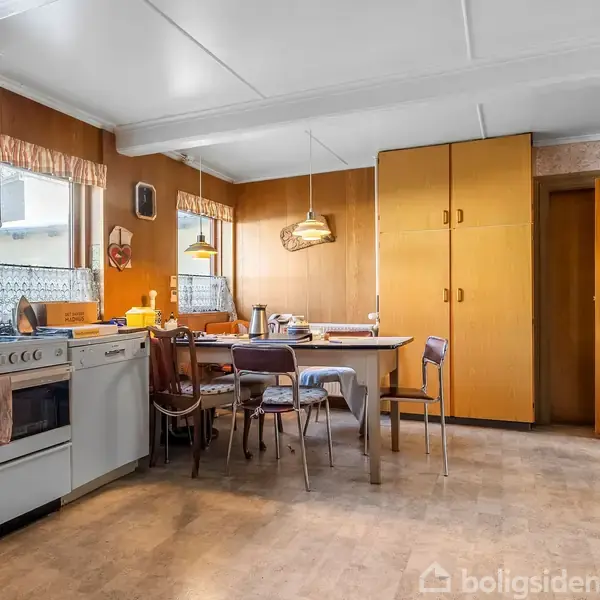 Køkken med træskabe og -paneler, spisebord med stole og en komfur- og ovnsektion til venstre; rummet er lyst op af loftslamper.