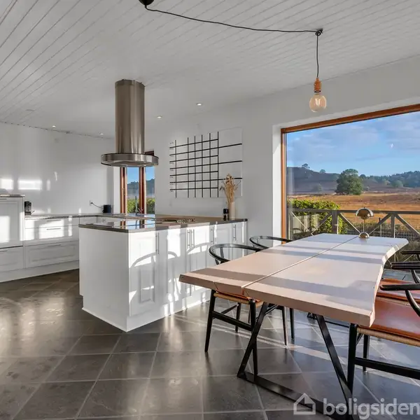 Køkken med hvidt interiør og centralt køkkenø. Et stort vindue viser udsigt over landligt landskab, mens et spisebord med stole står ved vinduet. En enkelt hængelampe ses ovenover.