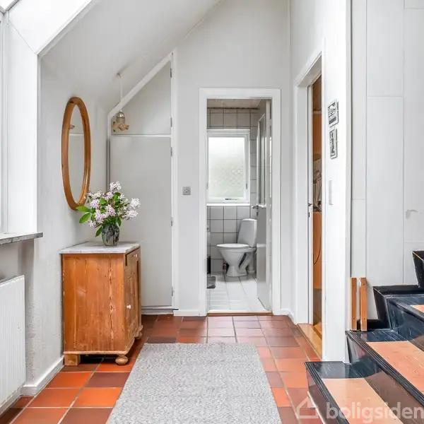 Entré med flisegulv og tæppe, indrettet med et skab og skænk med planter og blomster. Et rundt spejl hænger på væggen. I baggrunden ses et flisebelagt badeværelse.