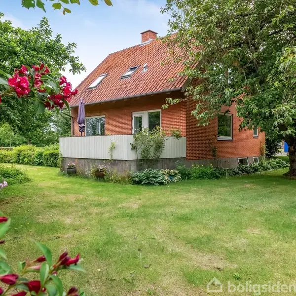 Rødt murstenshus med skråt tag står i en grøn have. Der er blomstrende buske og træer omkring, en terrasse med parasol og klart himmel.