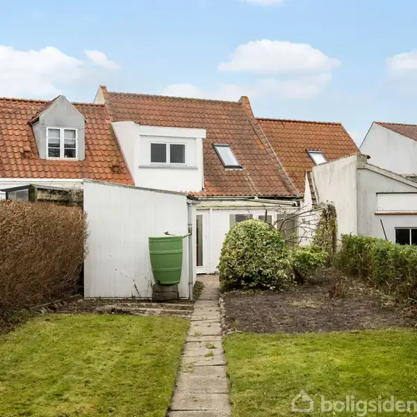 Hus med rødt tegltag og hvide mure står for enden af en smal stensti. Græsplæne og buske omgiver stien i en lille have.