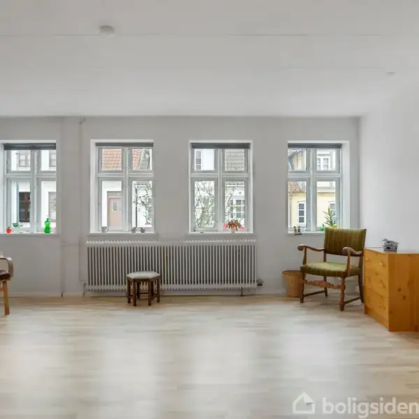 Stue med trægulv har to stole og en kommode foran tre store vinduer, der giver naturligt lys. Reoler fyldt med bøger står langs væggen.