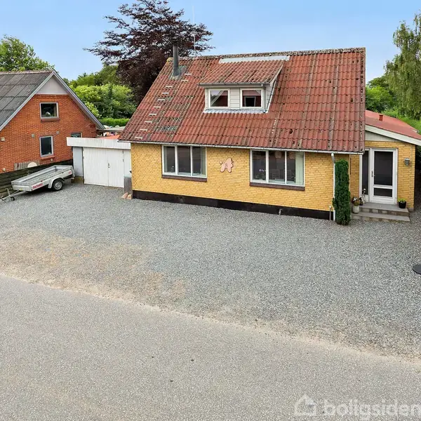 Gult murstenshus med rødt tegltag og hvidt garageport, står stille på grusplads. Omgivet af grønne træer og buske, en trailer til venstre. Klar blå himmel.