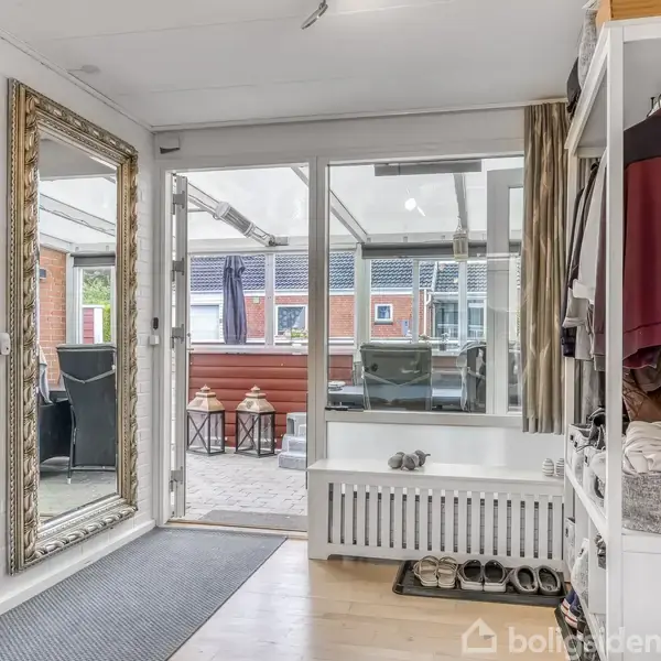 Et lyst indgangsparti med et stort gulvspejl til venstre, en åben garderobe med jakker til højre og en dør til en overdækket terrasse i baggrunden.