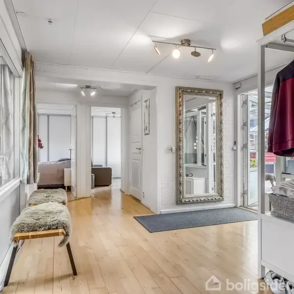 Entré med trægulv og et vægspejl. En bænk med fåreskind står til venstre, mens en åben garderobe med tøj er til højre. Åbne døre fører til andre rum.