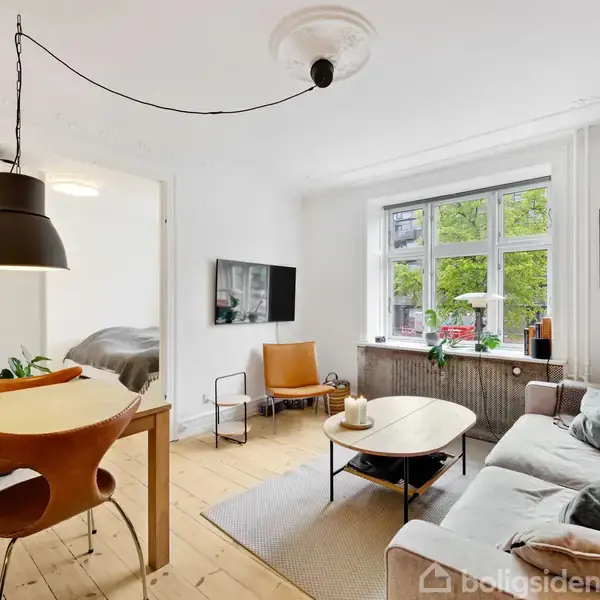 Stue med sofa og sofabord foran et vindue. Værelsesdør til venstre viser sengen. Trægulve, moderne indretning med kunst på væggen og grønne planter ved vindueskarmen.