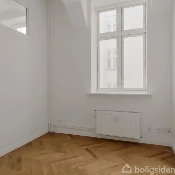 Et funktionelt køkken med grå skabe og en indbygget ovn. Rummet har hvide vægge og loft med indbygget belysning. Åben indgang til tilstødende rum.