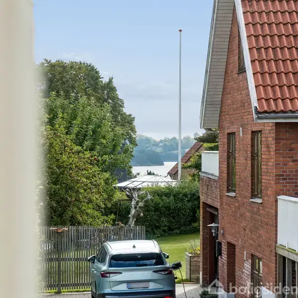 Biler holder parkeret foran et rødt murstenshus med rødt tegltag. I baggrunden ses en have med træer og udsigt til vandet. Atmosfæren er rolig og landlig.