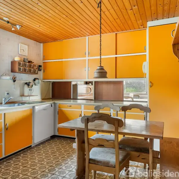 En køkken med orange skabe og hvide kanter. Et træbord med to stole står centralt. Lampe hænger over bordet. Kontekst inkluderer et vindue og mønstrede gulvfliser.