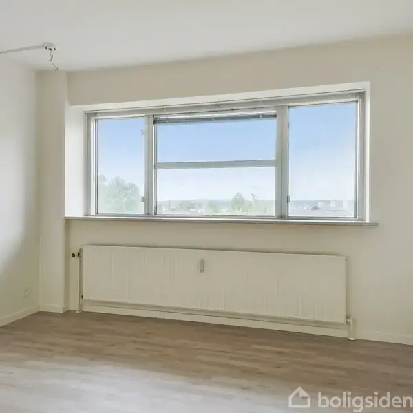 Et tomt rum har et stort vindue med udsigt til en klar himmel. Der er en radiator under vinduet og trægulv i rummet.