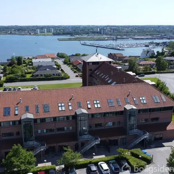 Bygning med rødt tegltag og udendørs trapper, set fra luften. Den ligger i et beboelsesområde nær en kystlinje med udsigt over en havn og by.