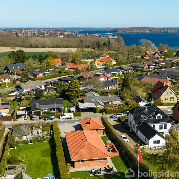 Huse med røde tage står i en række, omgivet af velplejede haver. En sø er synlig i horisonten, omkranset af træer og marker under en klar, blå himmel.