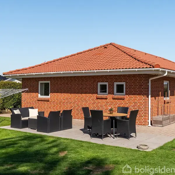 Rødt murstenshus med rødt tegltag. Terrasse med sorte havemøbler og en parasol i en grøn have. Klar blå himmel i baggrunden.