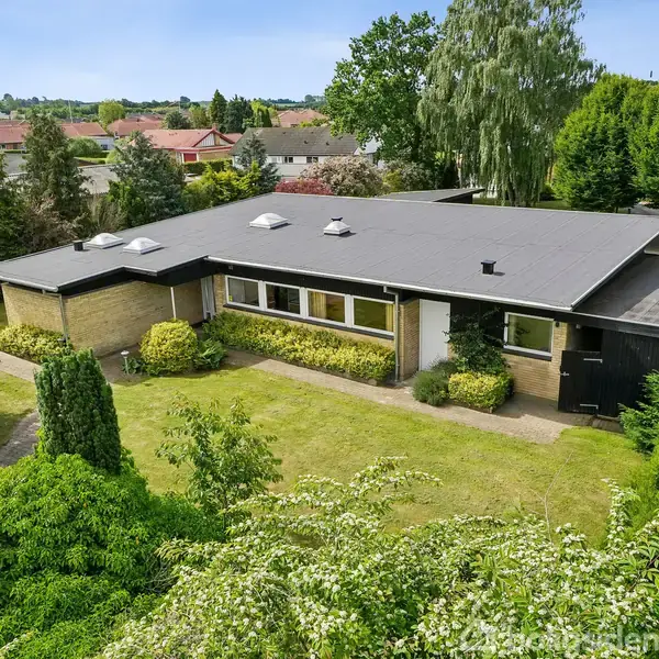 En flad tagstensvilla omgivet af velplejede buske og træer. Huset har et sort tag og store vinduer. Det ligger i et grønt boligområde med flere lignende huse i baggrunden.
